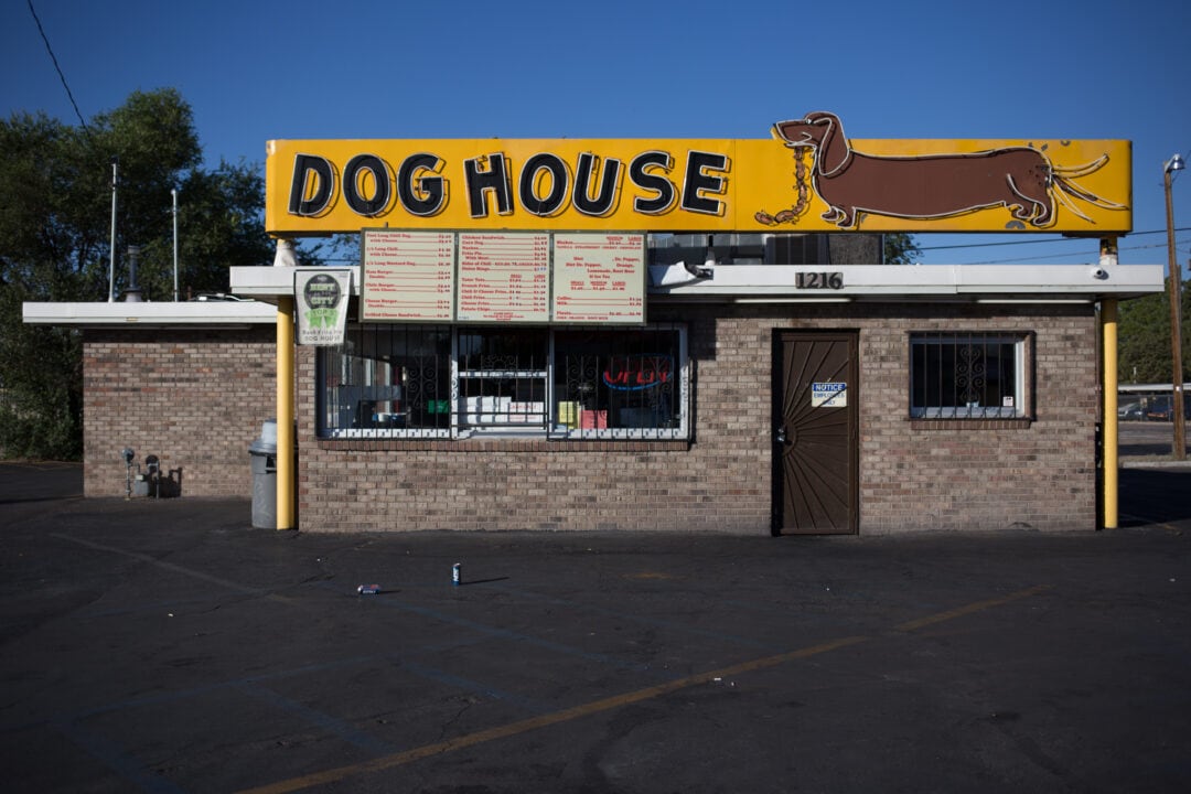 a small brick building topped with a large yellow neon sign featuring a dachshund and a string of sausages and the words "dog house"
