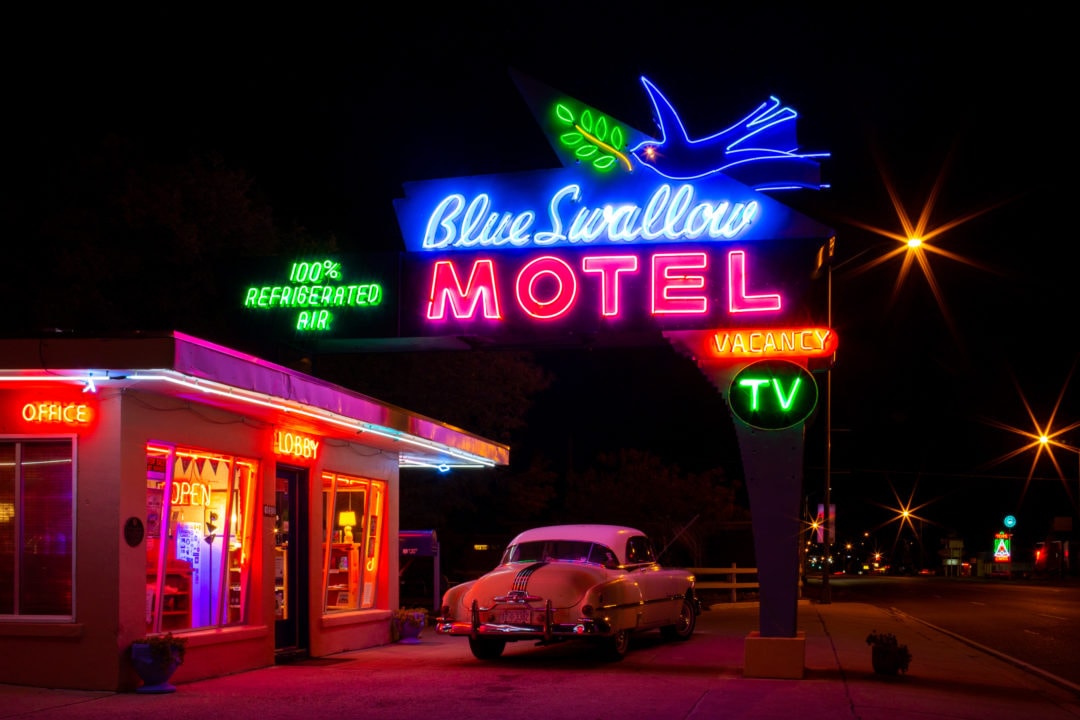 A vintage car is parked below a big neon sign that glows brightly in the dark night
