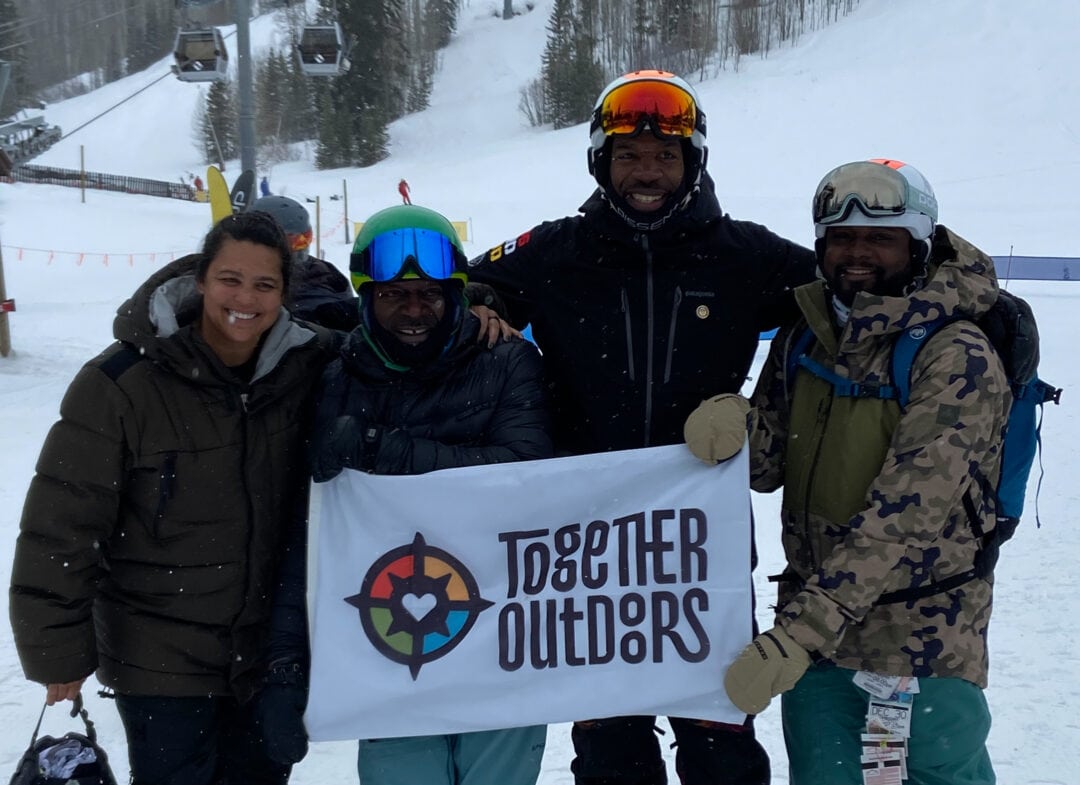 A group of winter adventurers hold up a sign that reads "Together Outdoors"