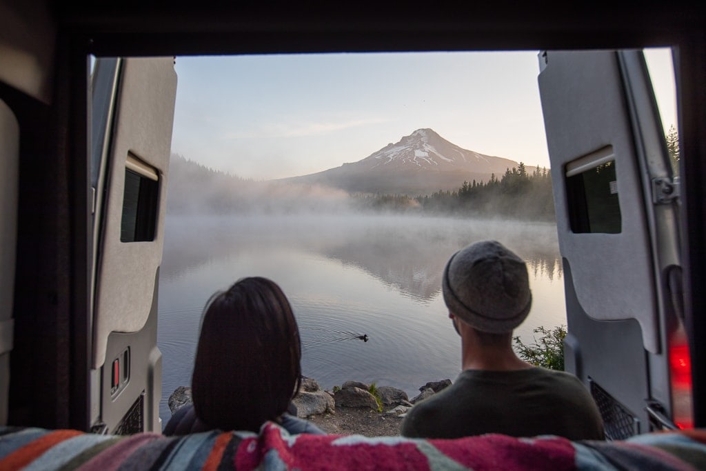 two people sit in the back of a campervan with the doors open looking at a lake with mountains in the background